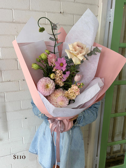 A hand-held bouquet of mixed wildflowers wrapped in pink paper and white tissue paper, held by a person wearing a light blue denim jacket in front of a white brick wall.