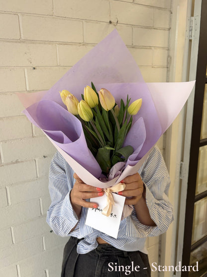A bouquet of yellow tulips wrapped in light purple and white paper, held by a person wearing a blue and white striped shirt in front of a white brick wall.