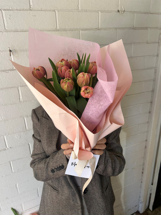 Person holding a bouquet of tulips wrapped in pink paper against a white tiled wall.