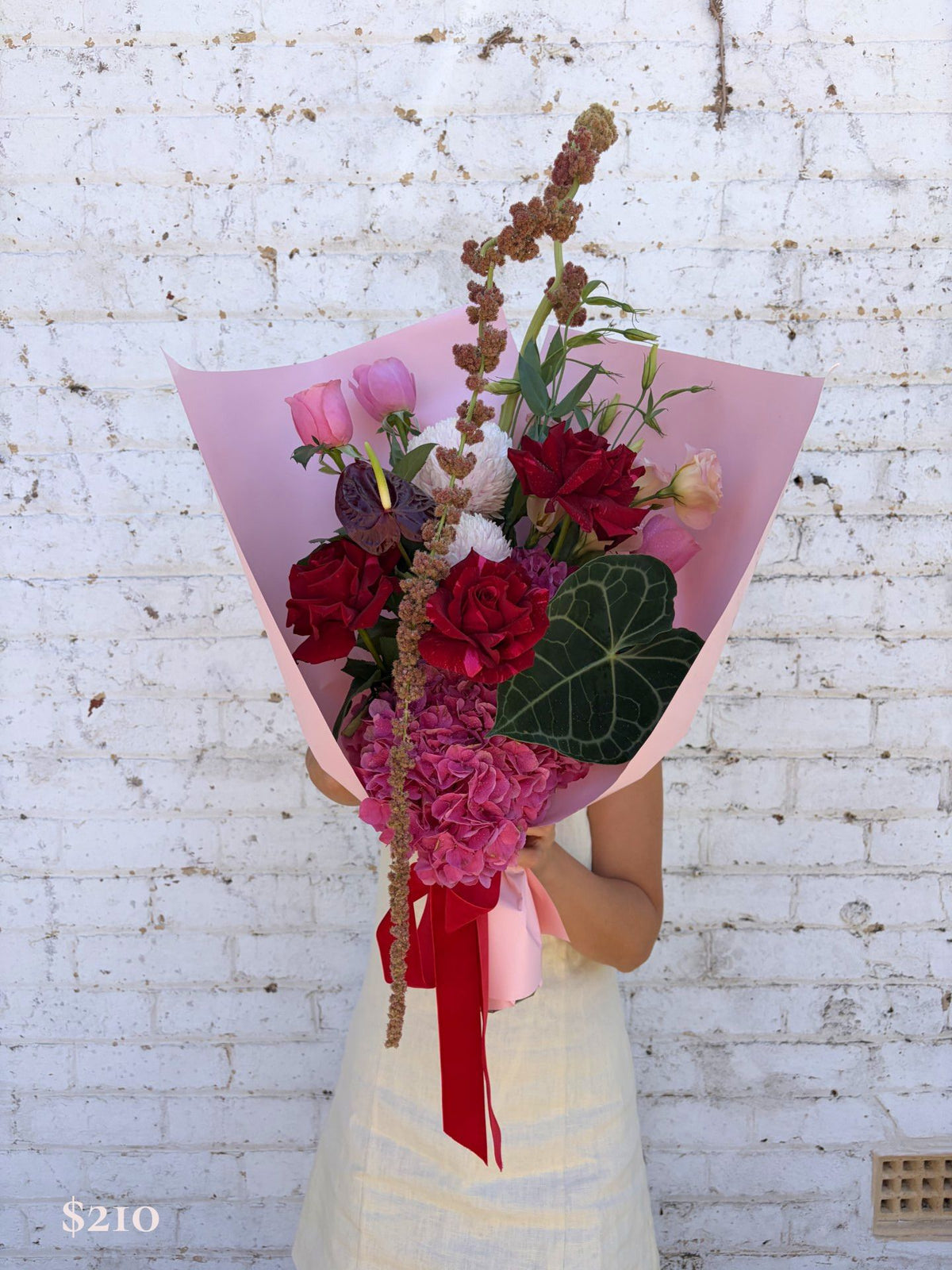 A person in a cream dress holds a large, vibrant flower bouquet wrapped in pink paper, featuring red roses, pink hydrangeas, and green foliage, set against a white brick wall.