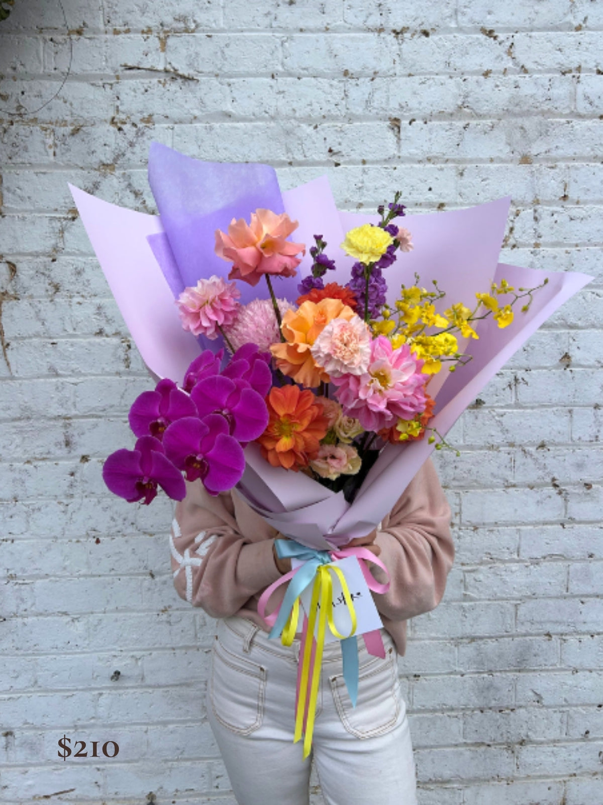 A person holding a large, vibrant flower bouquet wrapped in light purple paper, featuring purple orchids, orange and pink flowers, and yellow blossoms, set against a white brick wall.