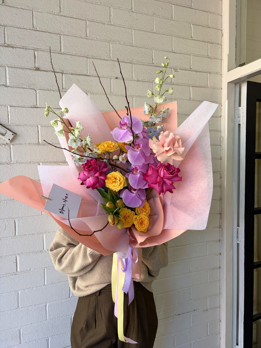 A person in a beige sweater holds a large, vibrant flower bouquet wrapped in pink and peach paper. The bouquet features purple orchids, yellow roses, pink carnations, and green foliage against a white brick wall background.