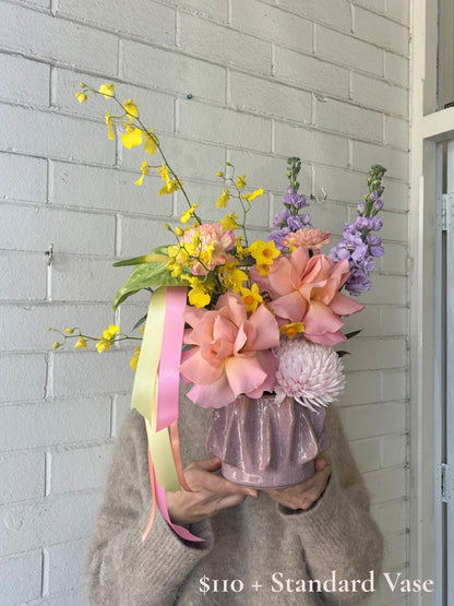 A person in a beige knit sweater holds a pastel flower arrangement in a shiny lavender-purple vase against a white painted brick wall. The bouquet includes pink flowers, yellow blossoms, and purple flowers, with pink and yellow ribbons hanging down.