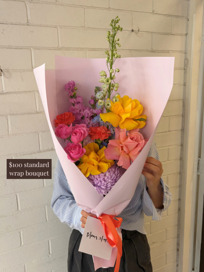 Bouquet of colorful flowers wrapped in pink paper held by a person against a light-colored wall.