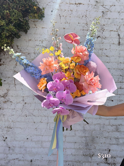 A vibrant floral bouquet wrapped in light purple paper, held by a person's arm against a white brick wall. The bouquet features purple orchids, peach roses, orange marigolds, blue delphiniums, and pink anthuriums, with pastel blue and yellow ribbons at the base.