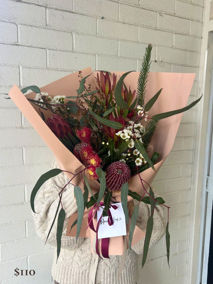 A hand-held floral bouquet wrapped in blush pink paper, featuring green eucalyptus leaves, burgundy protea-like flowers, red chrysanthemums, and small white flowers, with a cream card and ribbon. The bouquet is held by a person wearing a cream cable-knit sweater in front of a white painted brick wall.