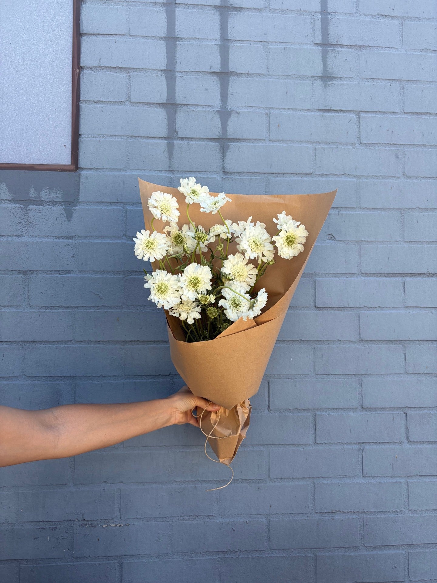 Bouquet of white flowers wrapped in brown paper held against a blue brick wall.