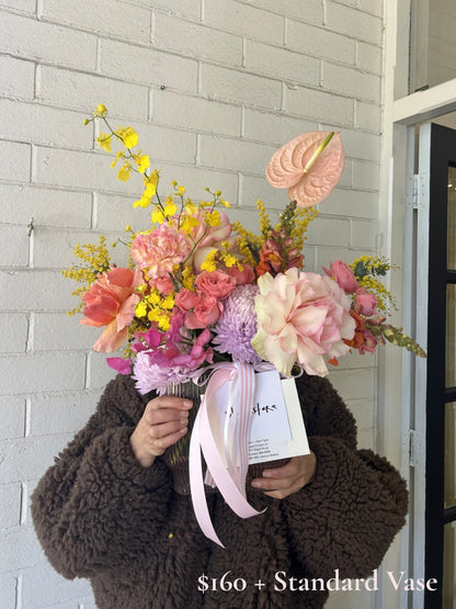A person in a brown fuzzy jacket holds a large, vibrant artificial flower bouquet with pink, yellow, and peach blossoms, set against a white brick wall.